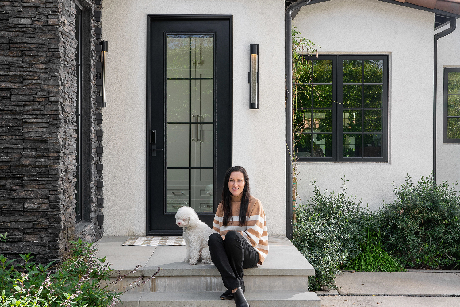 Laurie March sitting on the steps of her home with her white dog in front of a black entry door with glass slats in the middle.