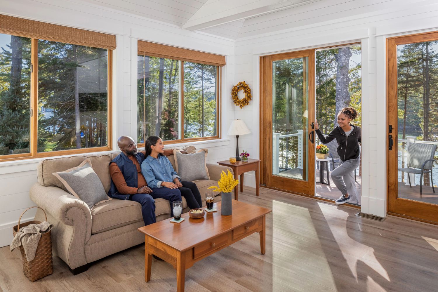 A brightly lit living room with windows all around and two sliding patio doors. All windows are framed in wood. There are two adults sitting on a couch and a child coming in the patio door.