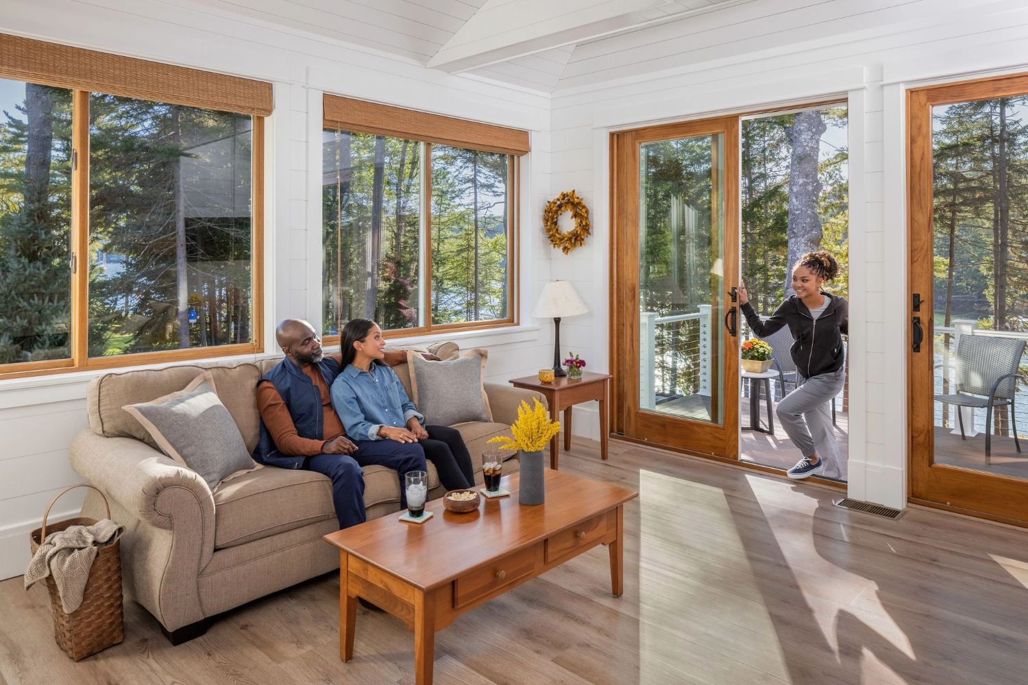 A brightly lit living room with windows all around and two sliding patio doors. All windows are framed in wood. There are two adults sitting on a couch and a child coming in the patio door.
