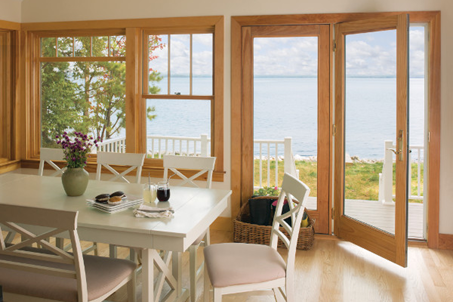 A natural wood finish french door that opens to a patio paired with a set of picture and casement windows in a dining room that look out over a body of water.