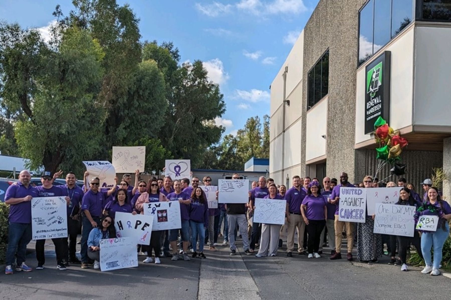 Group of RbA employees in purple t-shirts holding sigs with inspirational messages for United Against Cancer event.