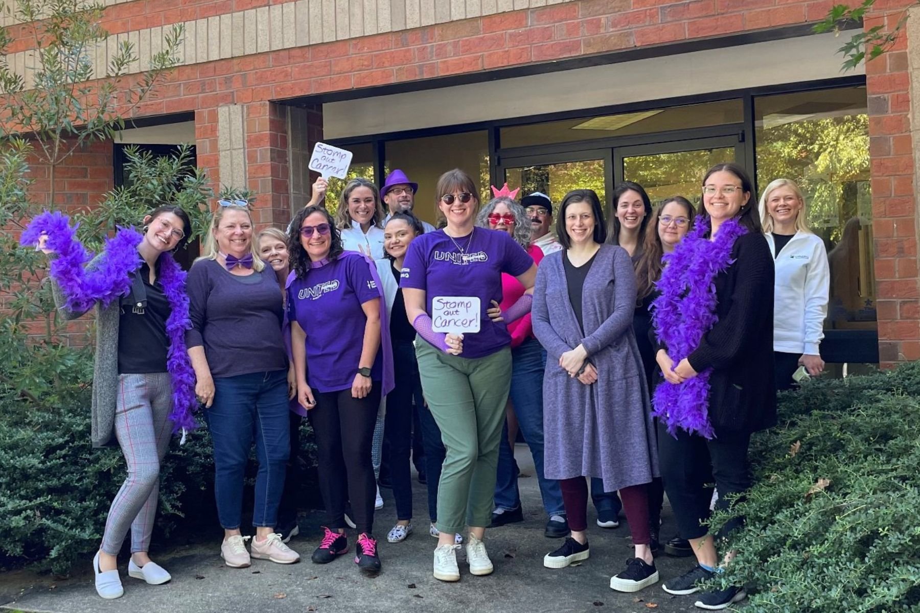 Group of RbA employees wearing purple t-shirts and costume, holding a sign that reads "Stomp oOut Cancer"