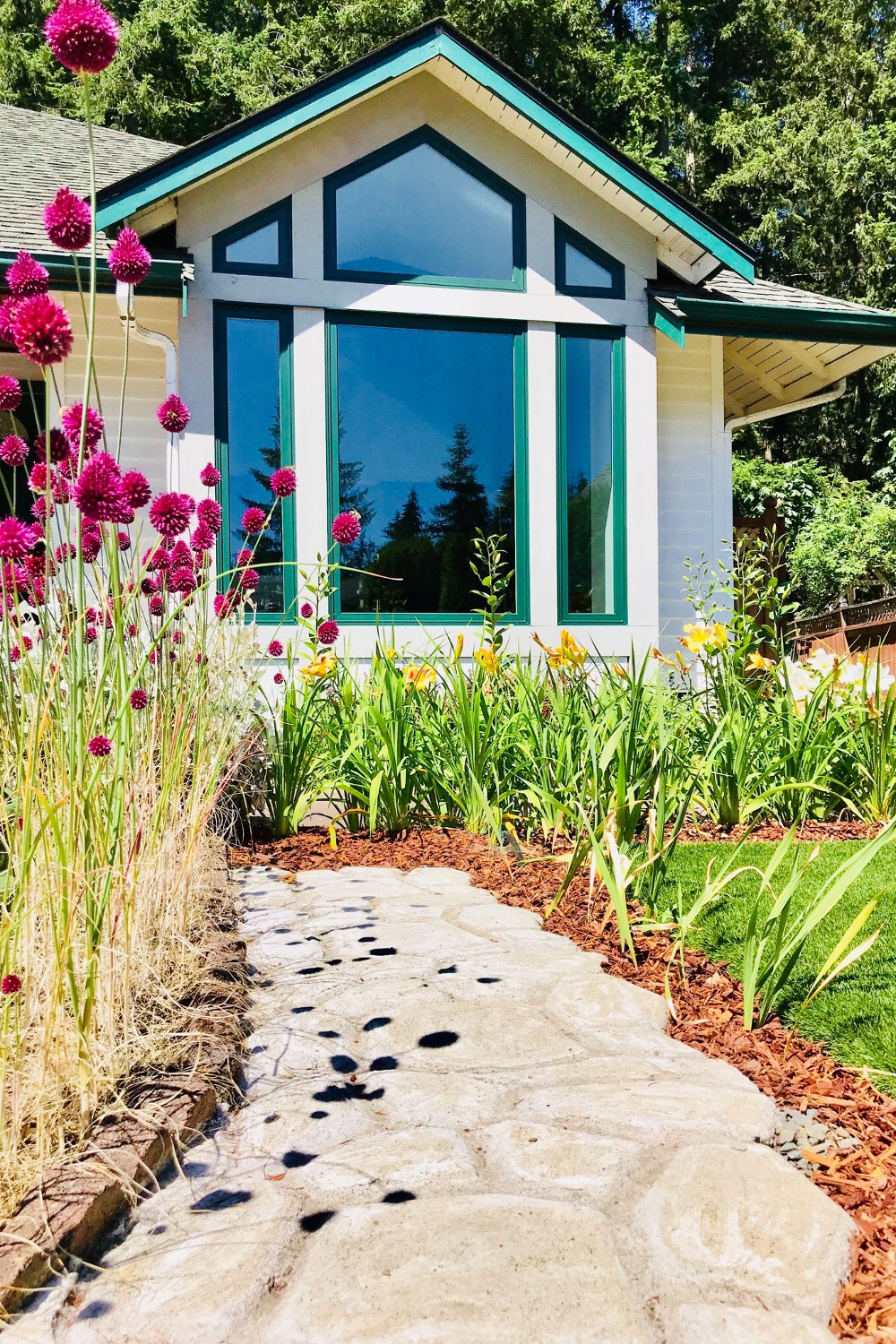 Two small trapezoid windows flank a peak pentagon window. These are set atop a bank of picture windows. The windows are forest green frames. There are pink flowers in the foreground providing contrast.