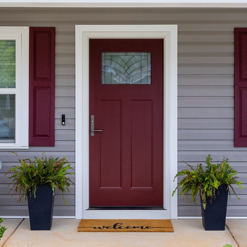 A single panel Ensemble entry door. The door has a small art glass panel. The door has white trim and brass hardware. It is red and matches the shutters.